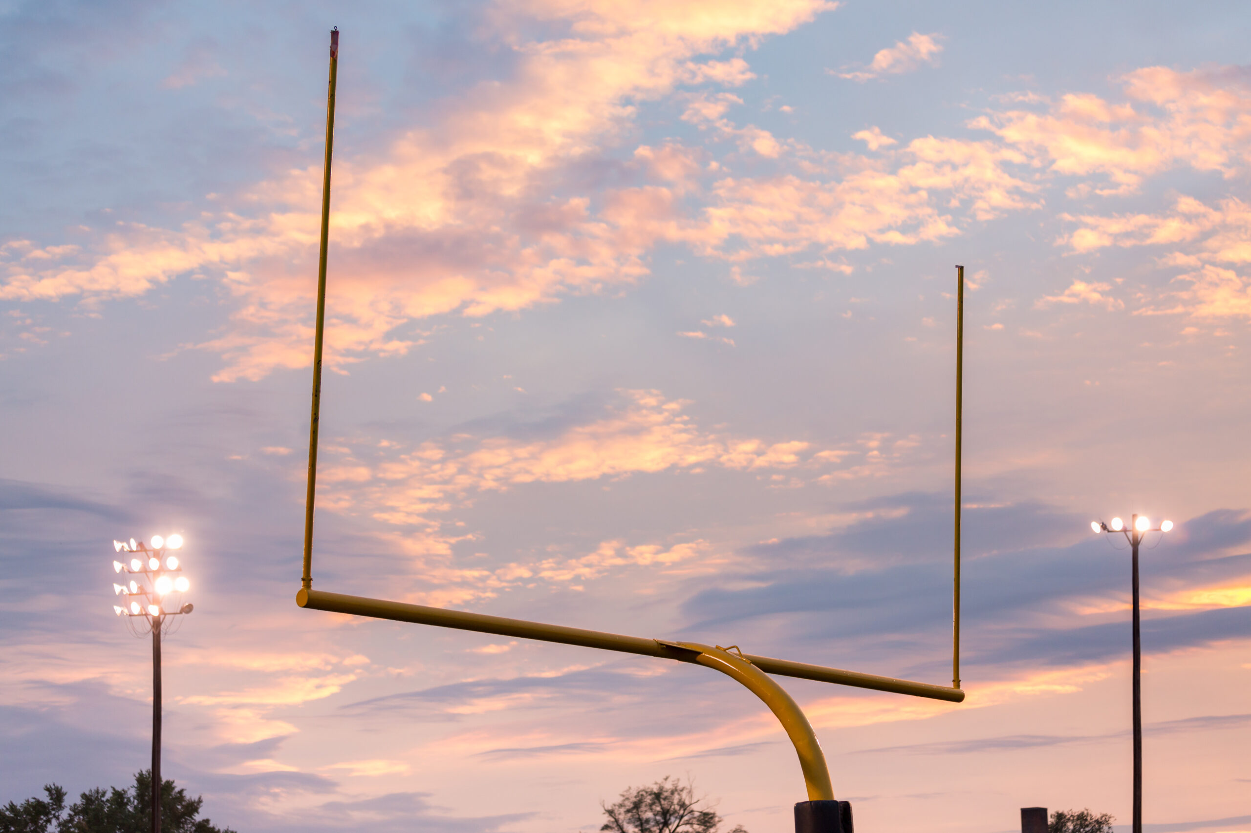 Yellow american football goal posts at school field against the lights and clouds of setting sun